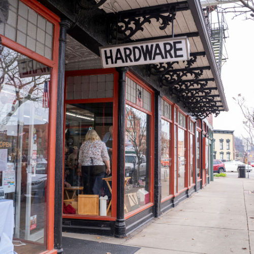 Vintage hardware store with red-framed windows and ornate black awning. A sign hangs above the entrance. Sidewalk and street visible in background.