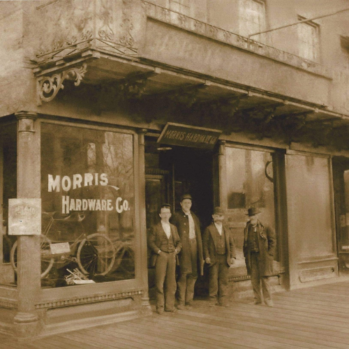 Sepia-toned photo of four men in period clothing standing outside the Morris Hardware Co. store. Display windows show bicycles; the setting evokes nostalgia.