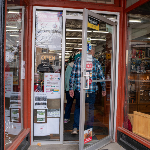 A person in a plaid shirt enters a hardware store through a glass door covered with posters and signs. The interior is warmly lit and inviting.