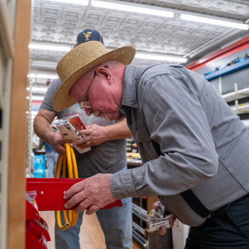 Older man in straw hat and glasses examines items in a hardware store. Another man in background holds tools, creating a focused, practical atmosphere.