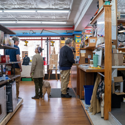A cozy store with wooden shelves filled with diverse gadgets and toys. Three people and a dog interact warmly at the counter, creating a welcoming atmosphere.