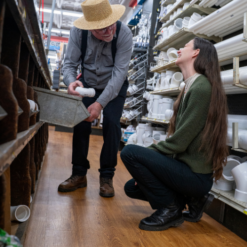An older man in a straw hat hands a PVC pipe to a seated woman with long hair in a hardware store aisle. They appear engaged and focused.