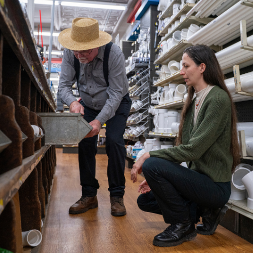 A man in a straw hat and a woman kneel in a hardware store aisle, examining a metal box. The shelves are stacked with pipes and fittings.