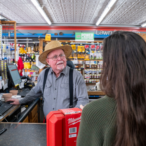 An older man in a straw hat and suspenders smiles behind a hardware store counter. A woman with long hair stands in front, holding a red toolbox. The setting is bright and inviting.